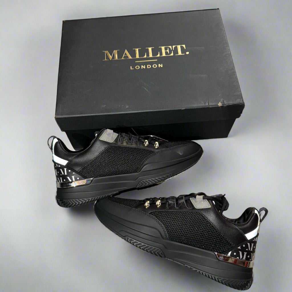 Black sneakers with gold accents on a concrete surface next to a black box labeled 'Mallet London'.