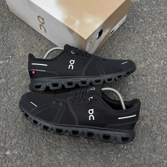 Black athletic shoes with 'on' branding on a gray surface, with a cardboard box in the background.