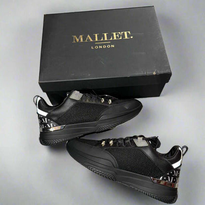 Black sneakers with gold accents on a concrete surface next to a black box labeled 'Mallet London'.