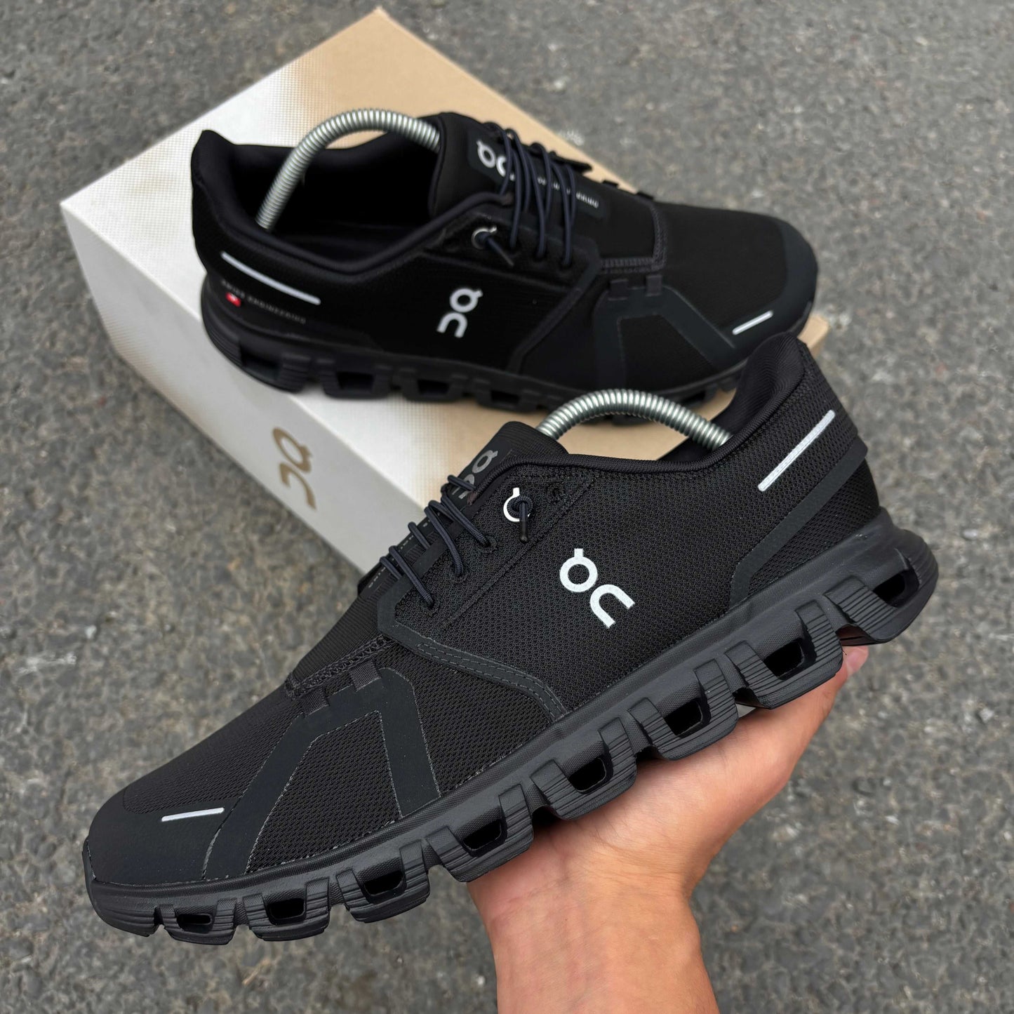 Black running shoes with unique sole design held by a hand, on a gray pavement background.
